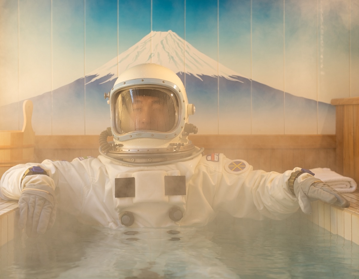 銭湯の湯船につかる宇宙飛行士。ヘルメットが湯気で曇り、背景に富士山のタイル絵がある写真